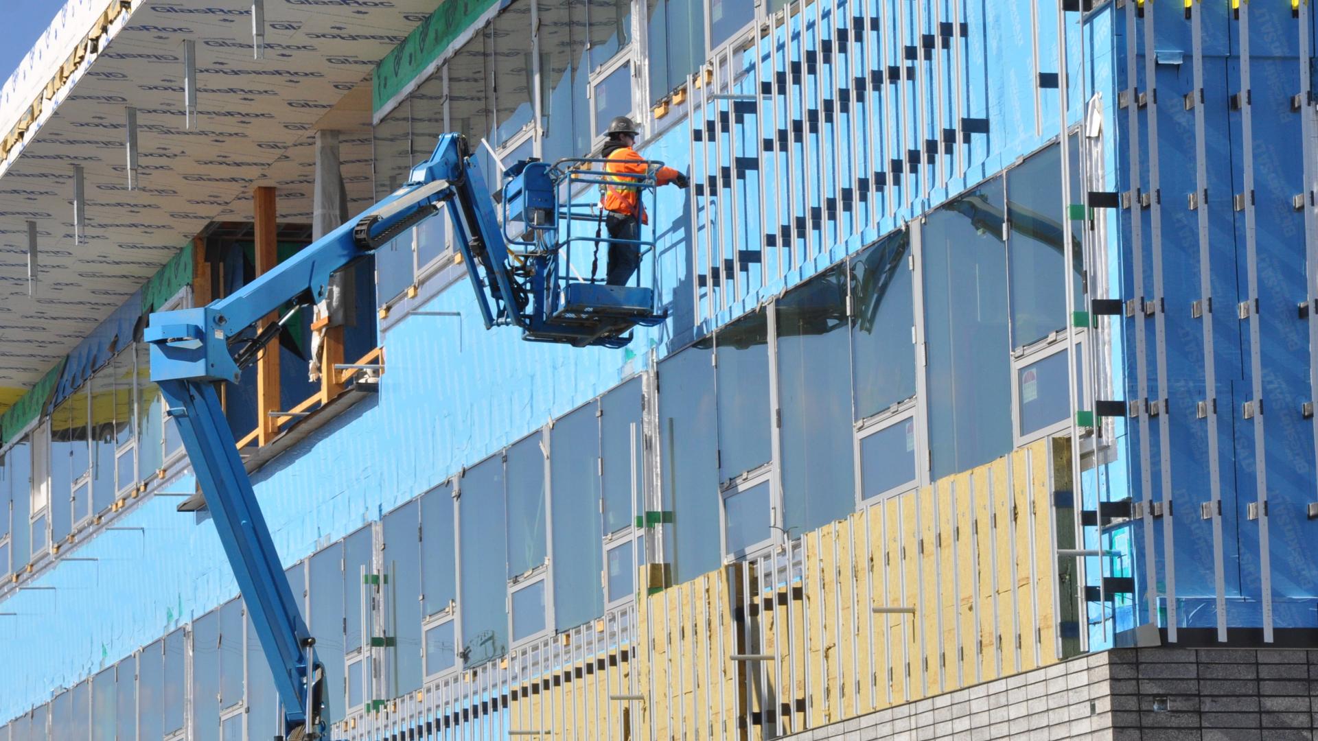 A construction worker in an orange vest on a blue lift installs siding on a building.