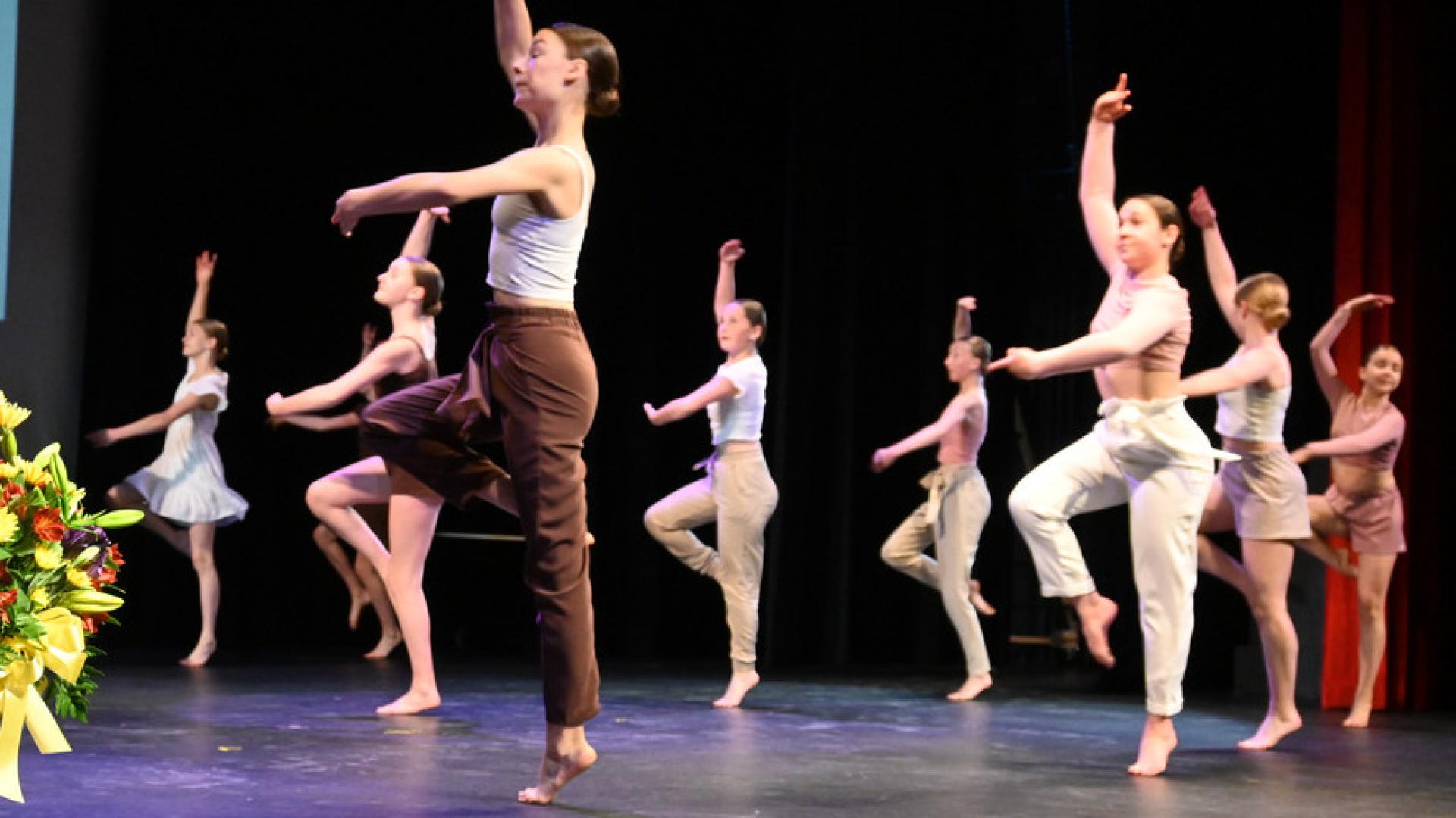 A group of dancers perform on stage, with one dancer in the foreground raising an arm and leg, while others mimic ballet poses in the background.