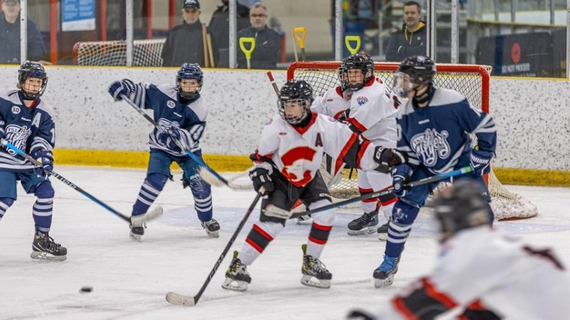 Youth hockey players in blue and white jerseys and red and white jerseys compete on the ice near the goal, with spectators watching in the background.