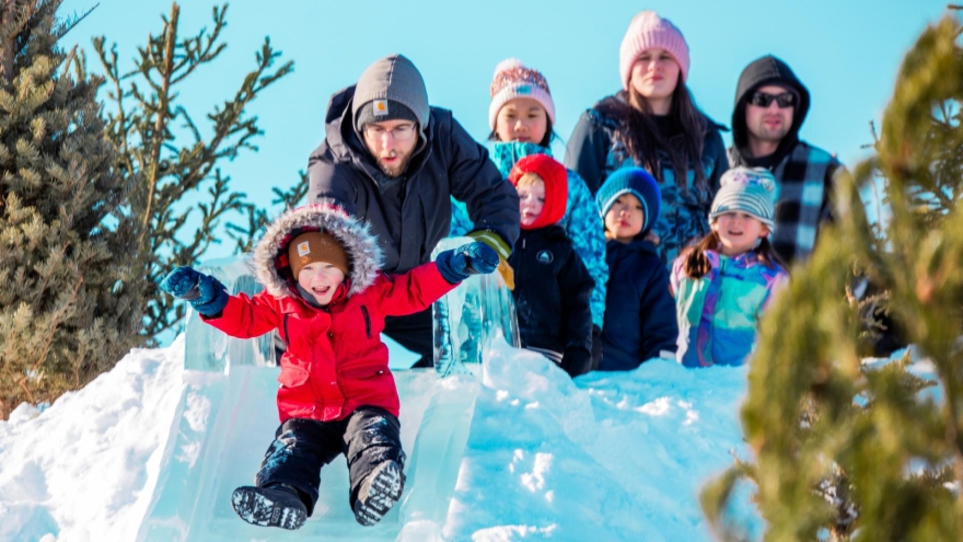 A child in a red coat excitedly sleds down an ice slide, surrounded by adults and children wearing winter apparel. 