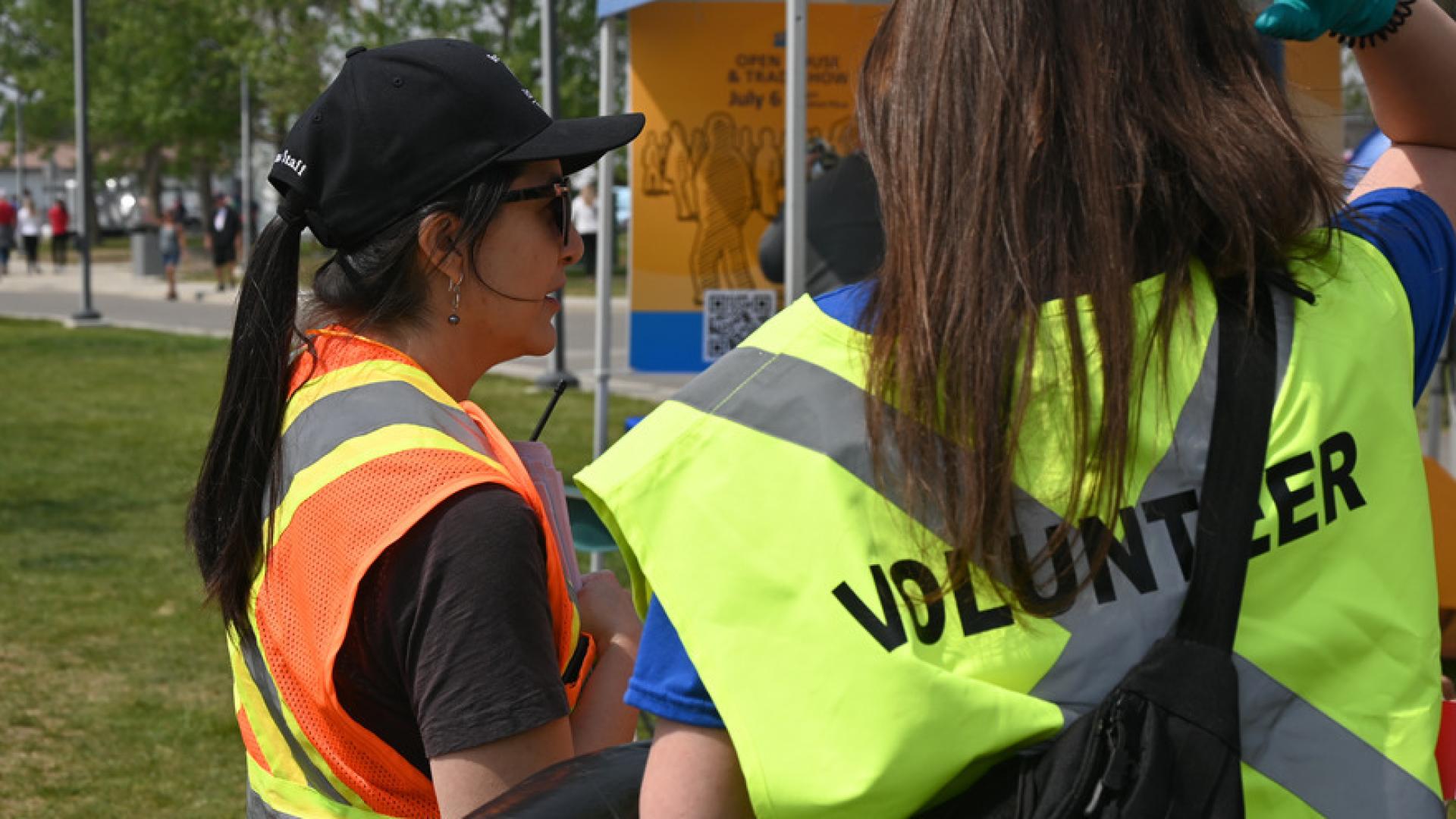 Two volunteers in safety vests and caps converse outdoors during an event.