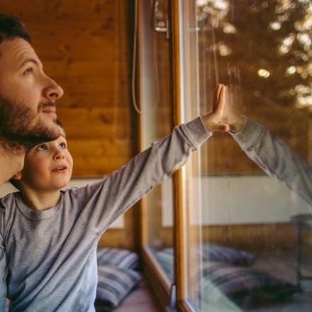 Person and child gaze out a window in a wooden cabin, with warm lighting.