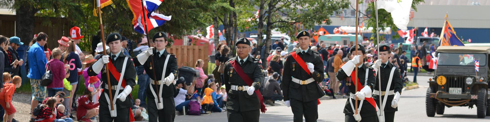 Cadets march in the annual Canada Day parade carrying flags down a city street lined with people.